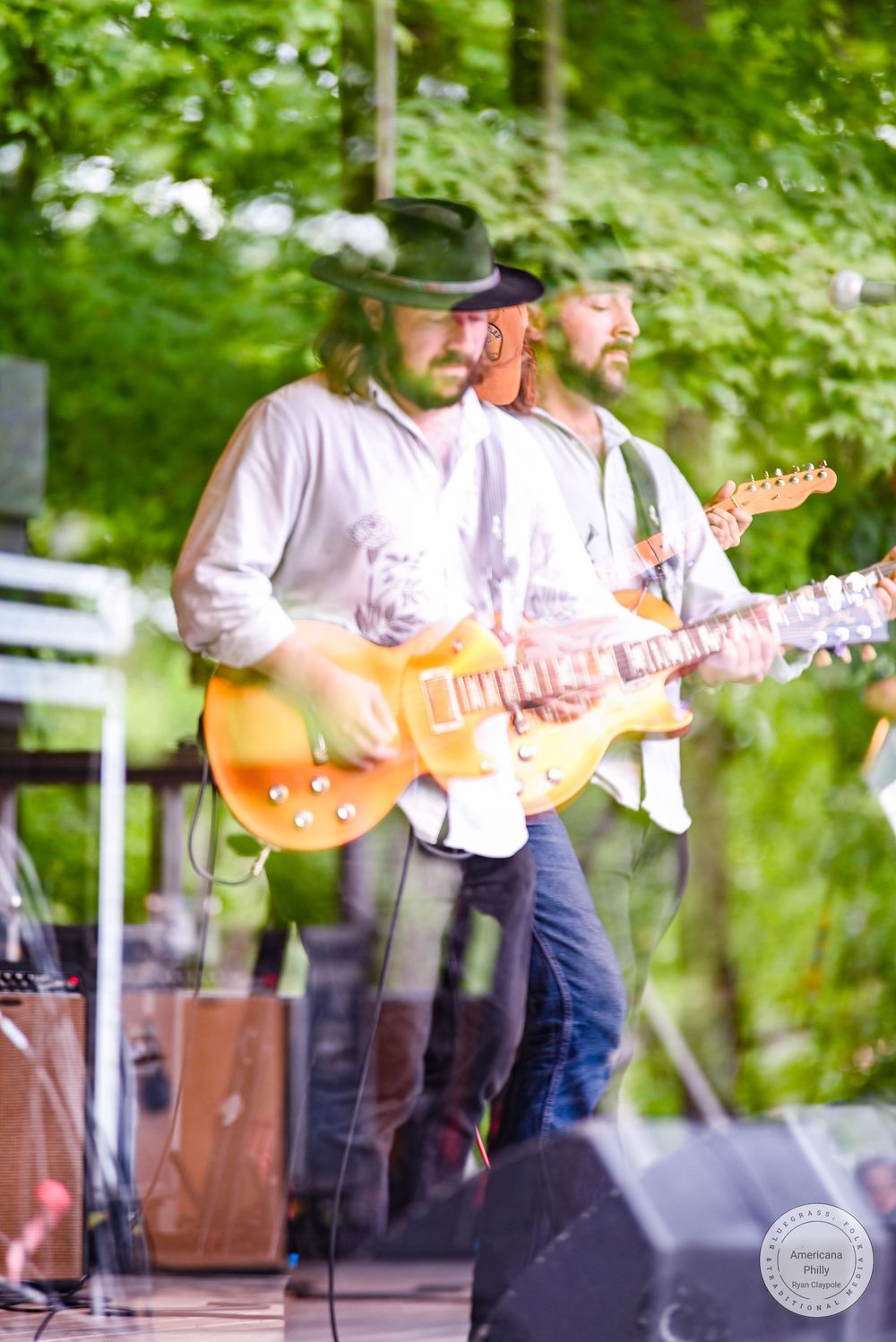 Two guitarists perform outdoors on stage surrounded by lush green trees, one wearing a black hat playing a gold electric guitar.