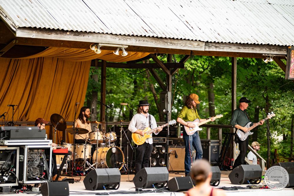 Live band performing on an outdoor stage with gold curtains, featuring guitarists and drummer at a summer festival.