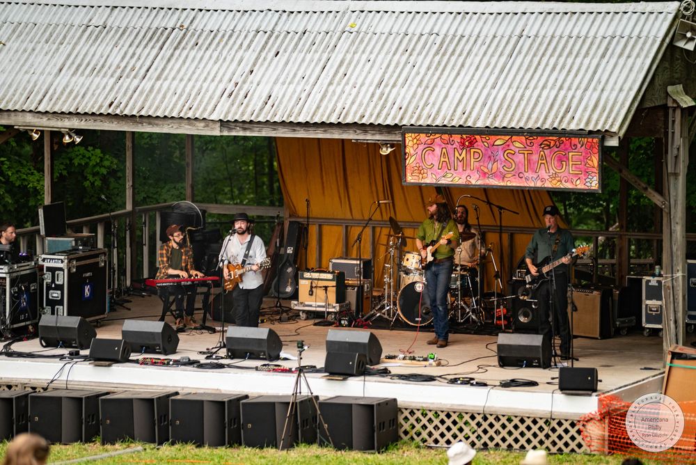 Band performing live on outdoor stage under metal roof at a sunny music festival.