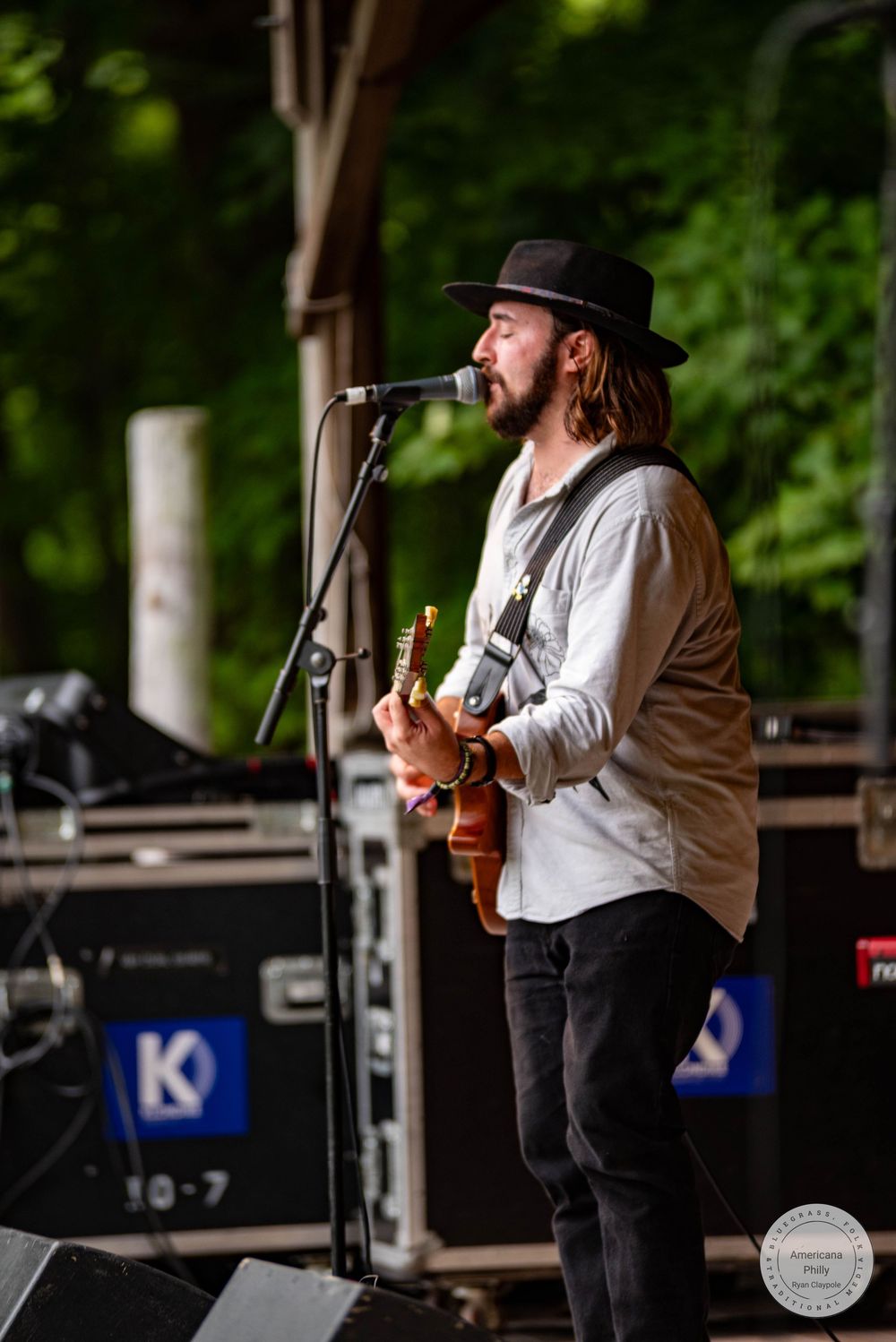 Male guitarist with long hair and black hat performs on outdoor stage with microphone, surrounded by lush green trees.