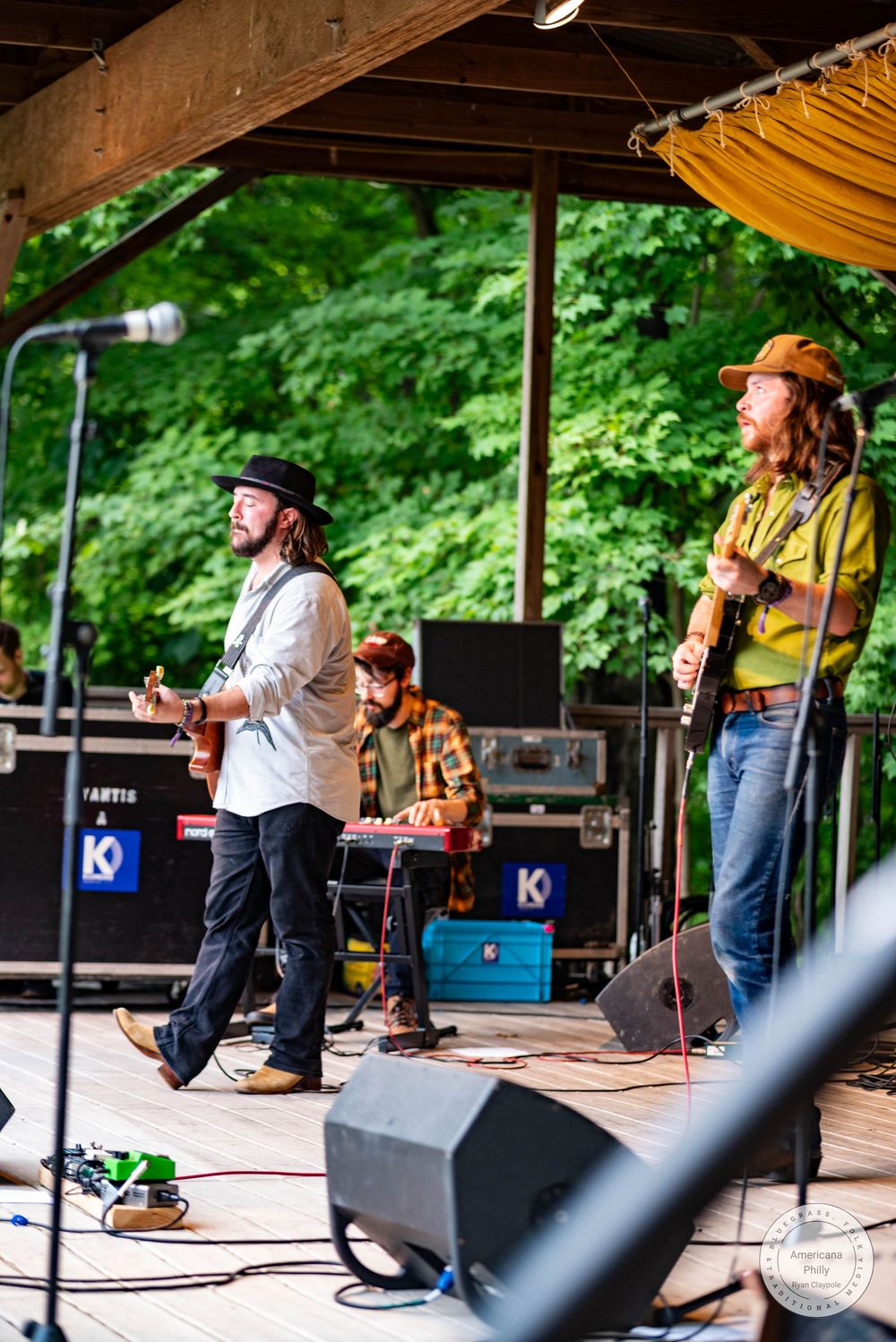 Three musicians perform on an outdoor stage surrounded by lush green trees at a summer music festival.