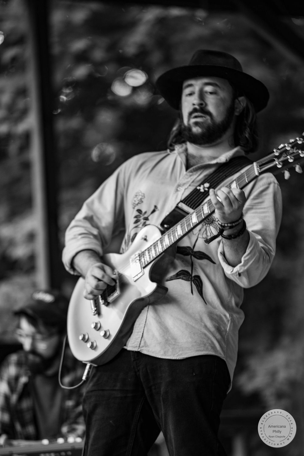 Black and white photo of bearded guitarist in wide-brim hat playing a Les Paul electric guitar on stage.