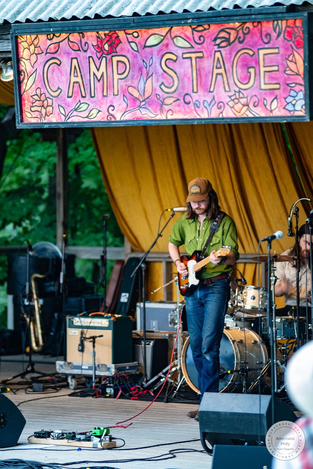 Guitarist performing on Camp Stage at outdoor music festival, wearing green shirt and jeans, with drums visible behind.