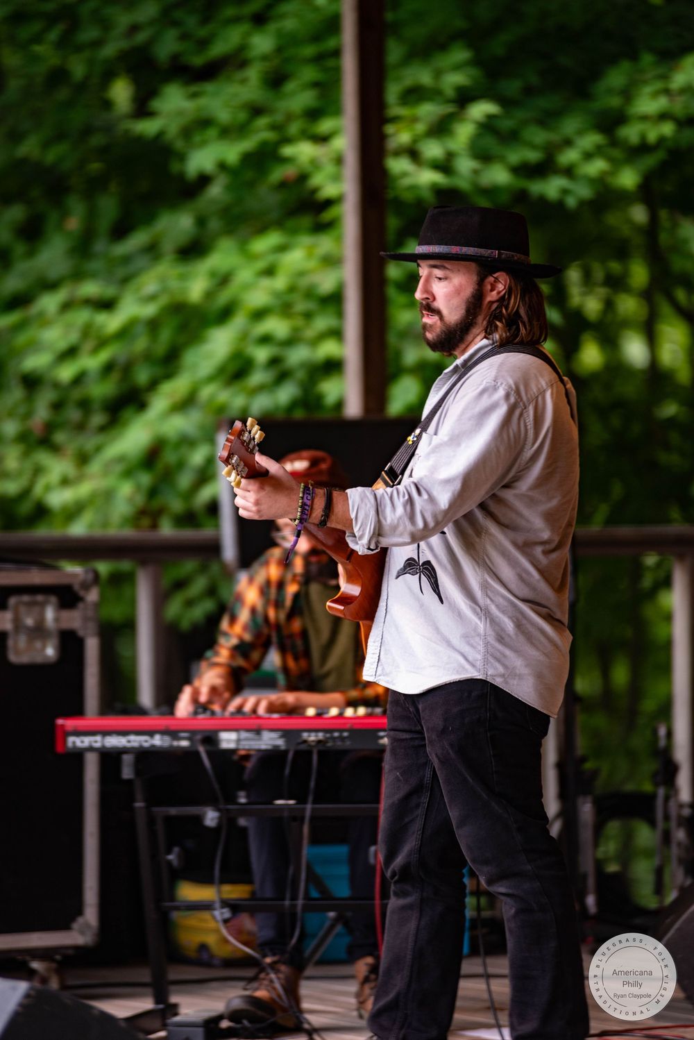 Bearded guitarist in black hat performs on outdoor stage with keyboardist and lush green trees in background.