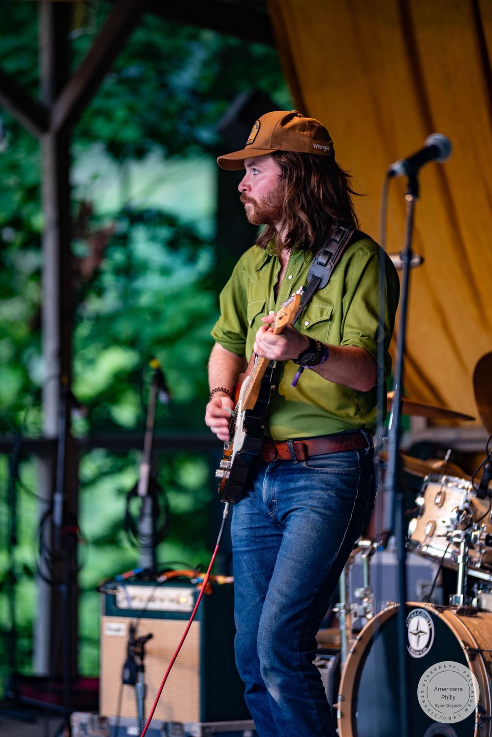 Musician in green shirt and brown cap plays electric guitar on outdoor stage at music festival.