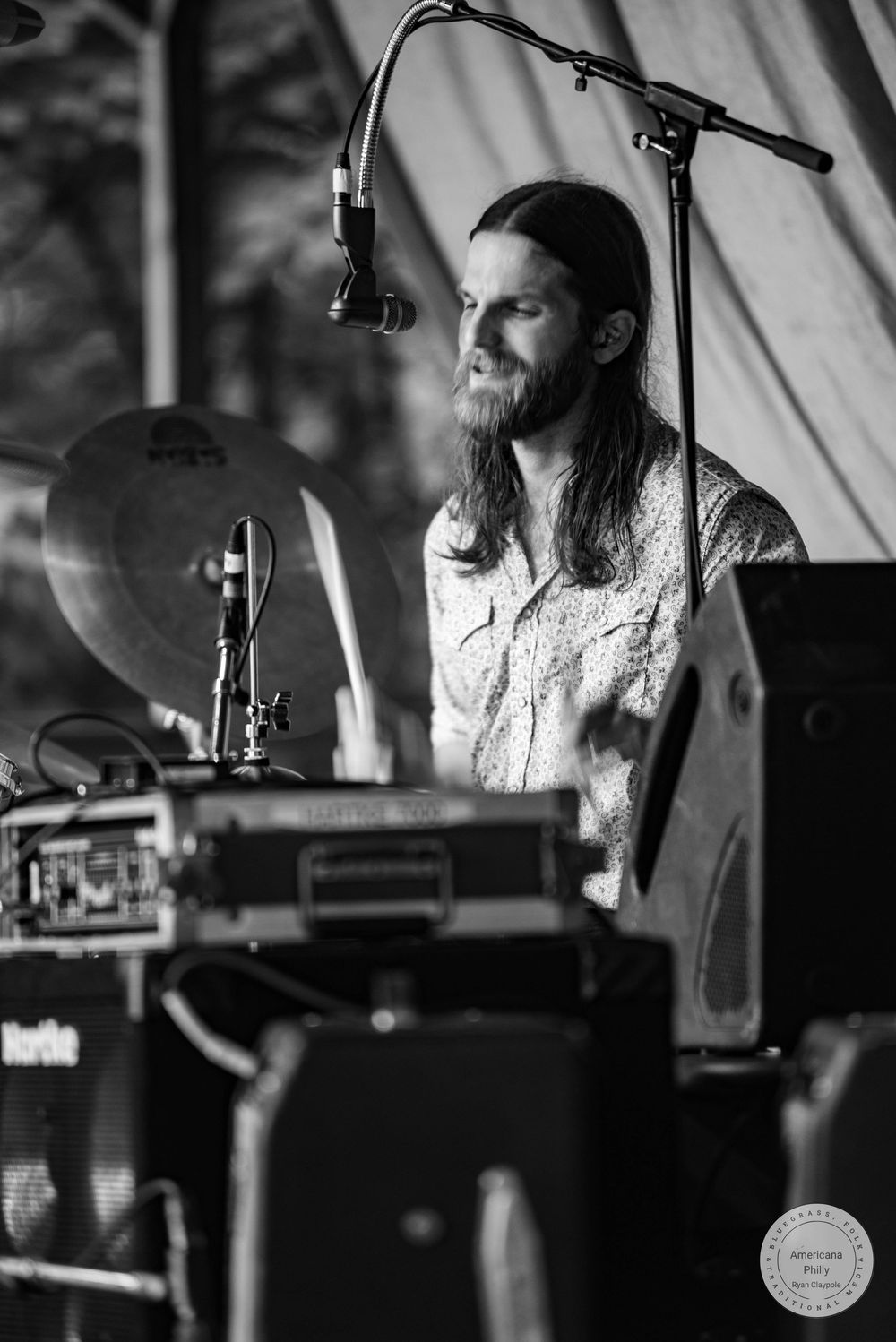 Black and white photo of a bearded musician with long hair performing on stage surrounded by drums and microphones.