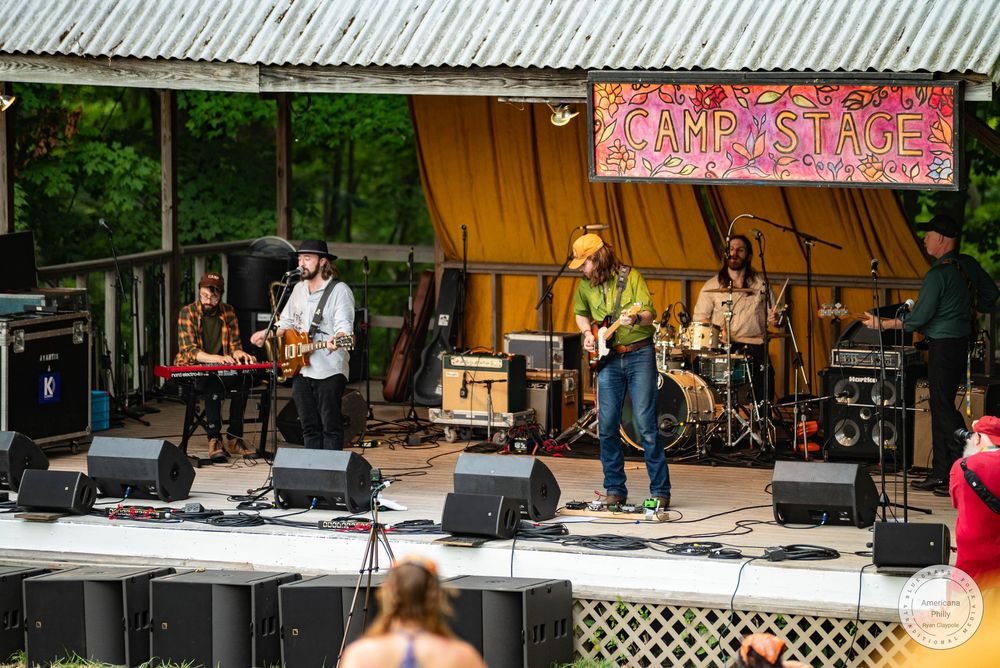 Live band performing on an outdoor stage at a music festival surrounded by lush green trees.
