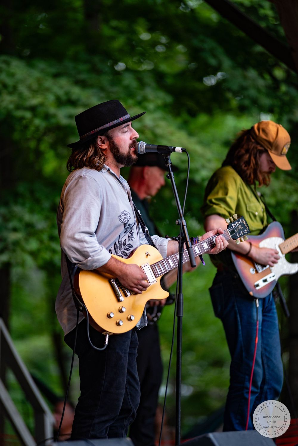 Bearded musician in black hat plays gold Les Paul guitar on outdoor stage with band members behind him.