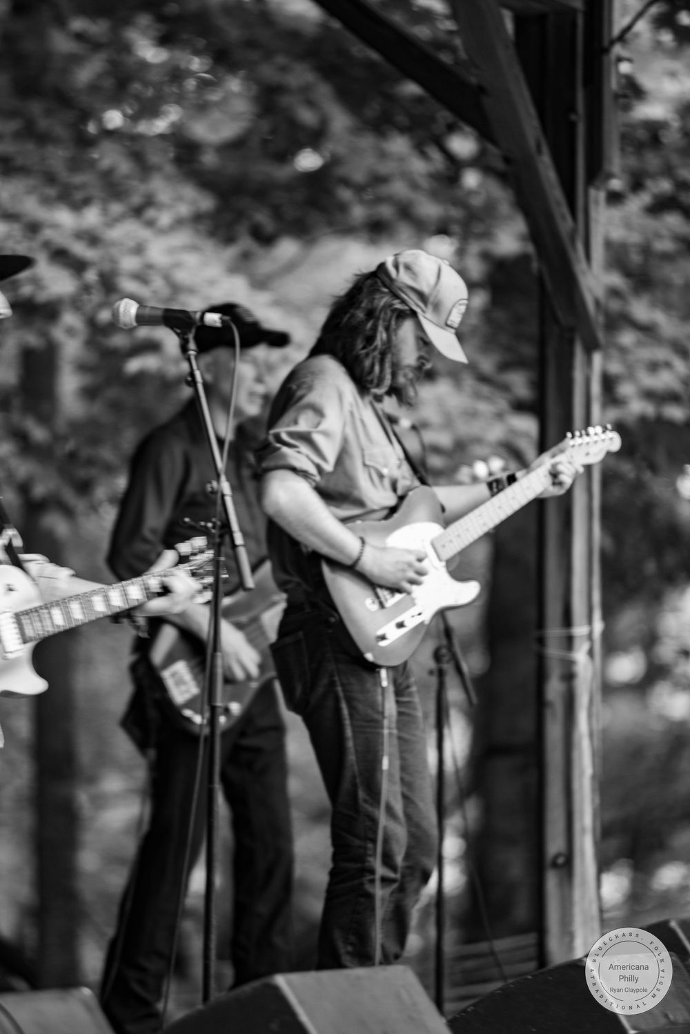 Black and white photo of a male guitarist performing on an outdoor stage, wearing a cap and playing an electric guitar.