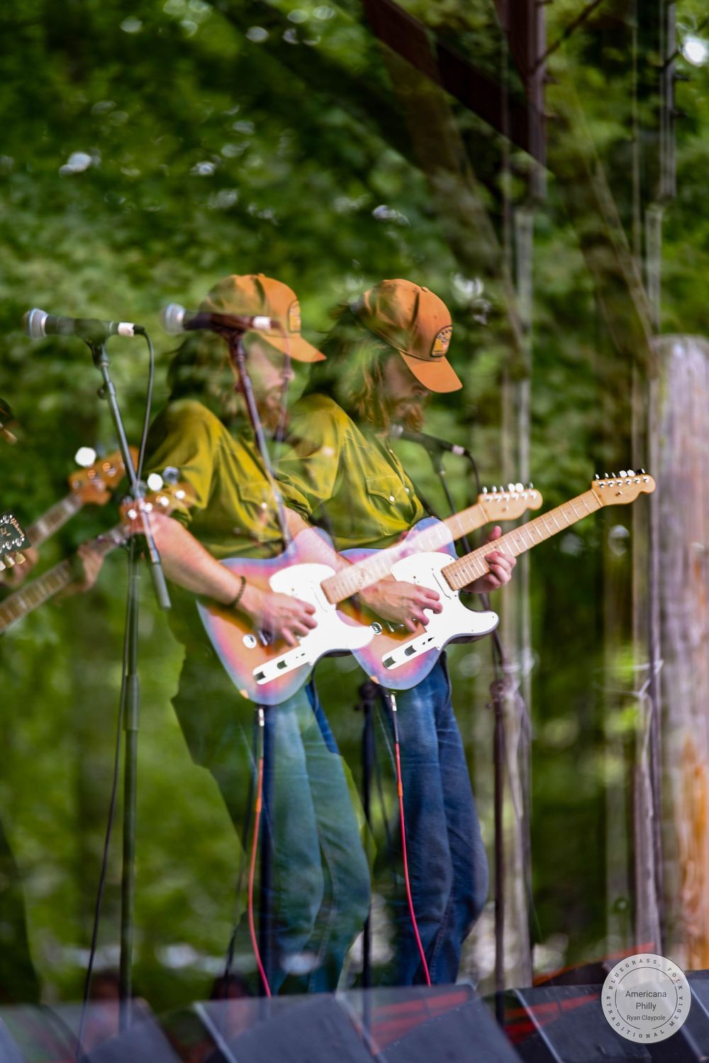 Guitarist in orange cap plays pink Stratocaster outdoors, reflected in glass at a live music festival.