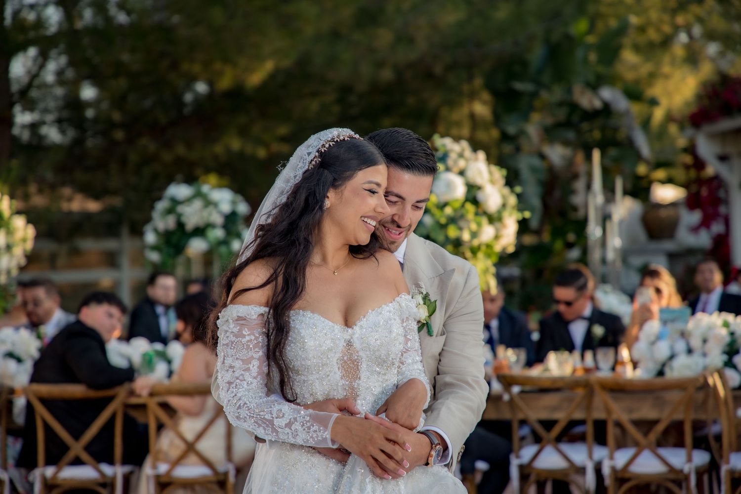 Bride and groom share a romantic embrace at an outdoor wedding reception, surrounded by guests and floral decorations.
