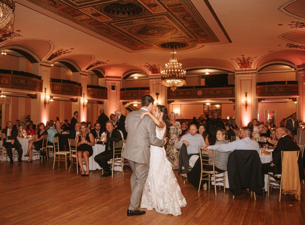 Bride and groom share first dance in elegant ballroom with ornate ceiling, warm lighting, and seated wedding guests.