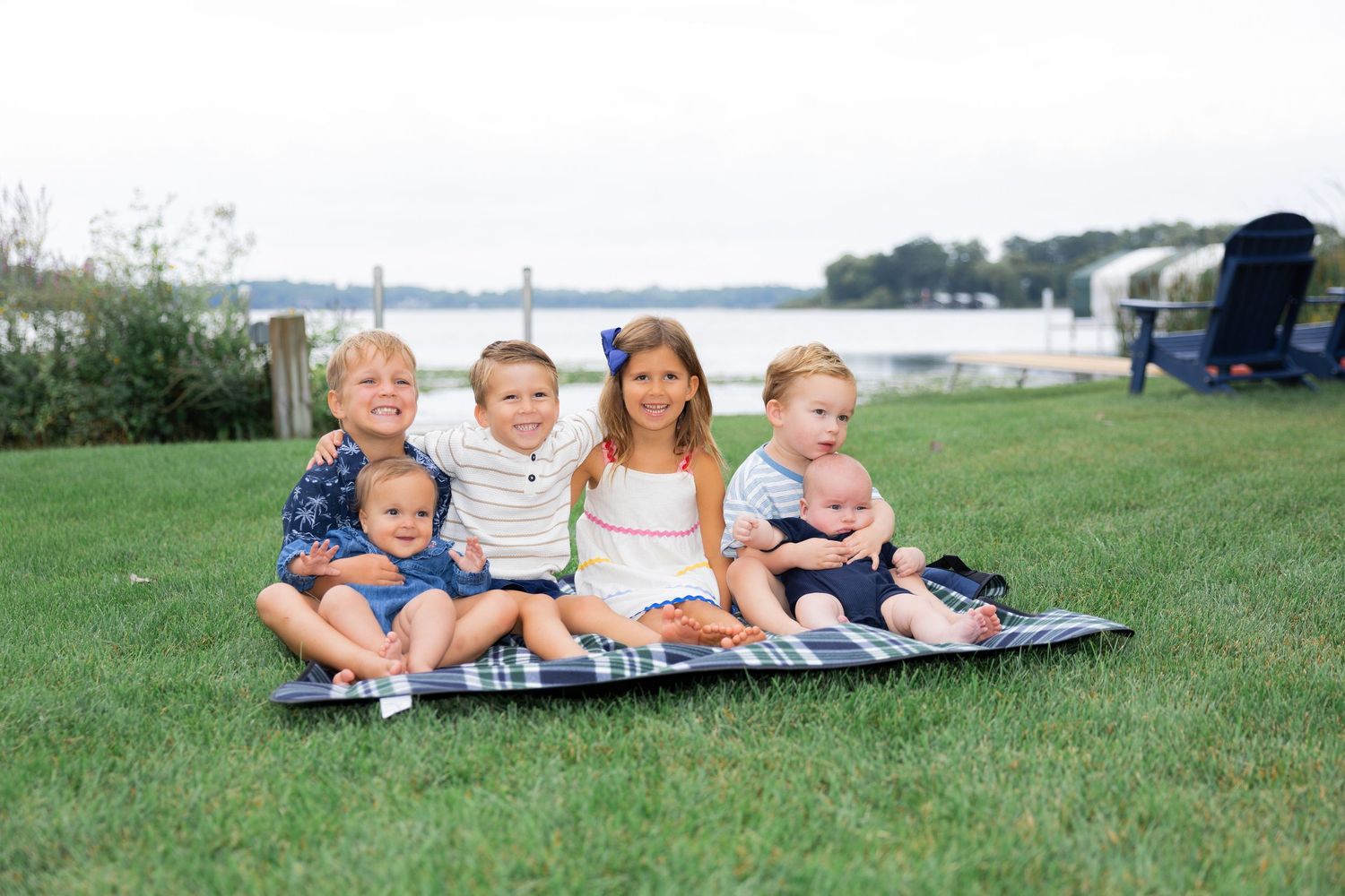 Six young children sitting together on a blanket on green grass near a waterfront on a cloudy day.