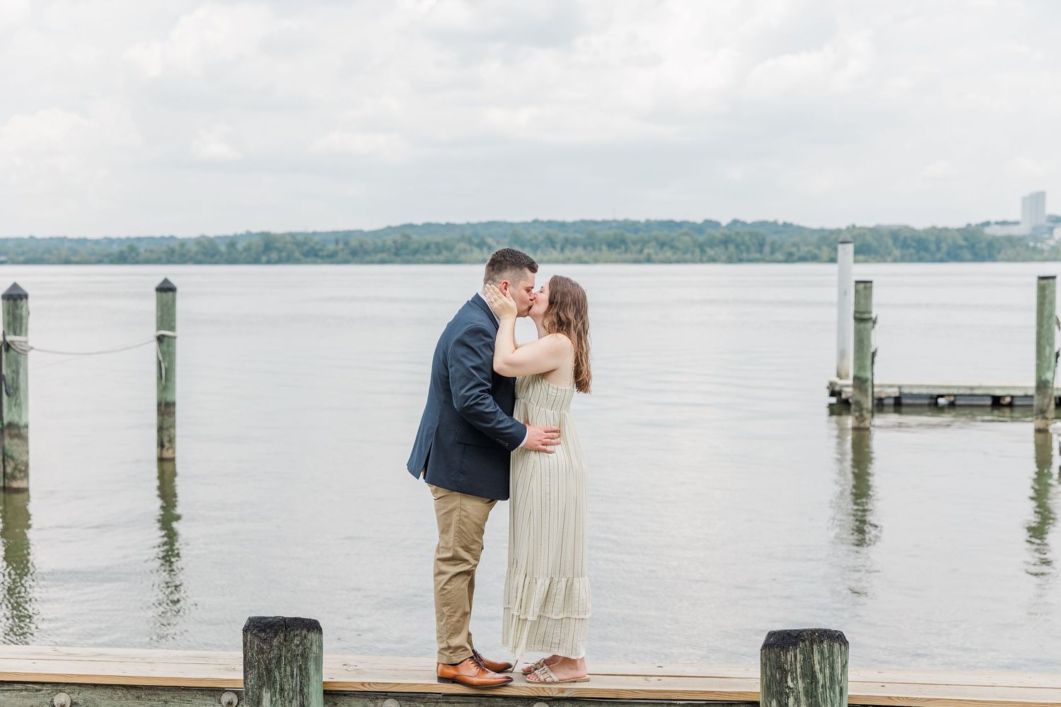 Couple sharing a romantic kiss on a wooden dock overlooking a calm river with lush green trees in the background.