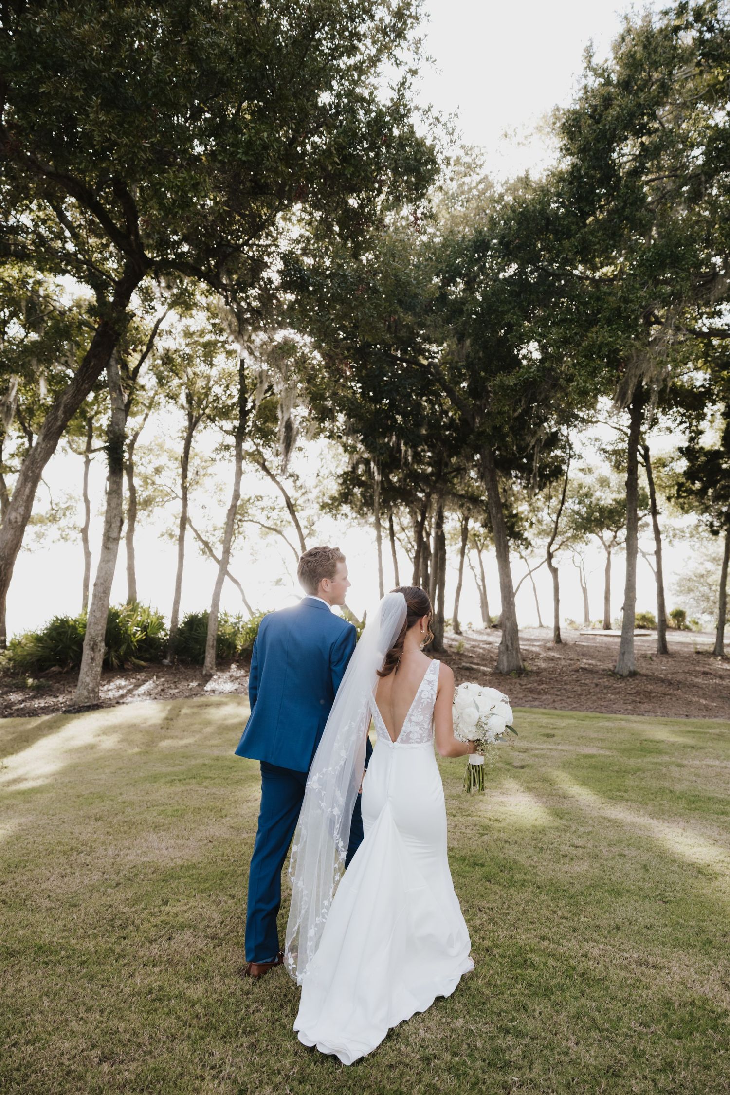 Bride and groom walking hand in hand through sunlit tree-lined lawn after outdoor wedding ceremony.