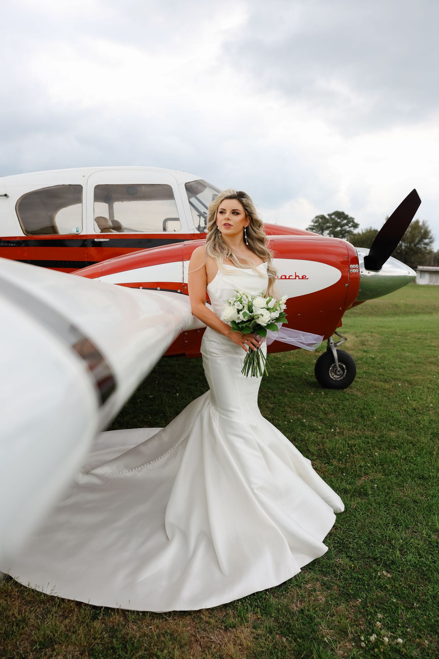 White wedding gown with flowing train photographed in front of a red and white private airplane on grassy field.