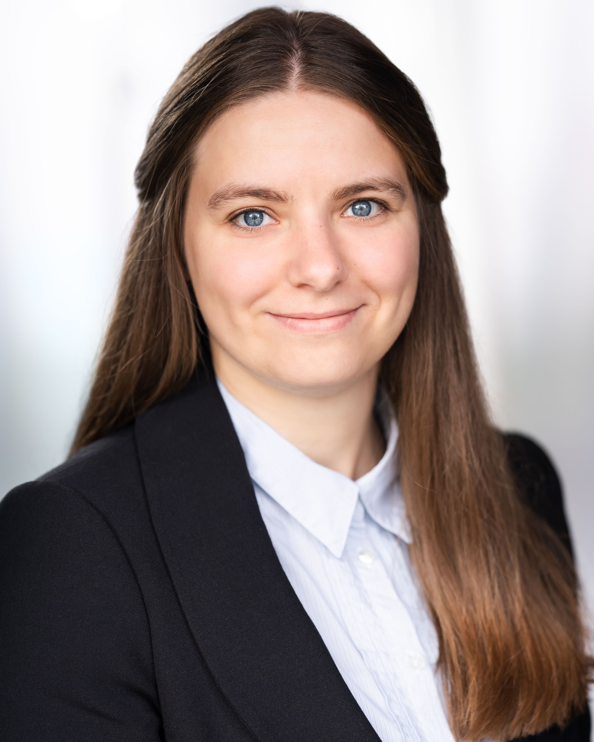 Professional headshot of someone in a dark blazer and white button-up shirt with long brown hair against a light background.