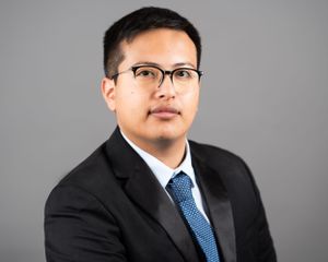 Professional headshot of a person wearing glasses, black suit and blue tie against a gray background.