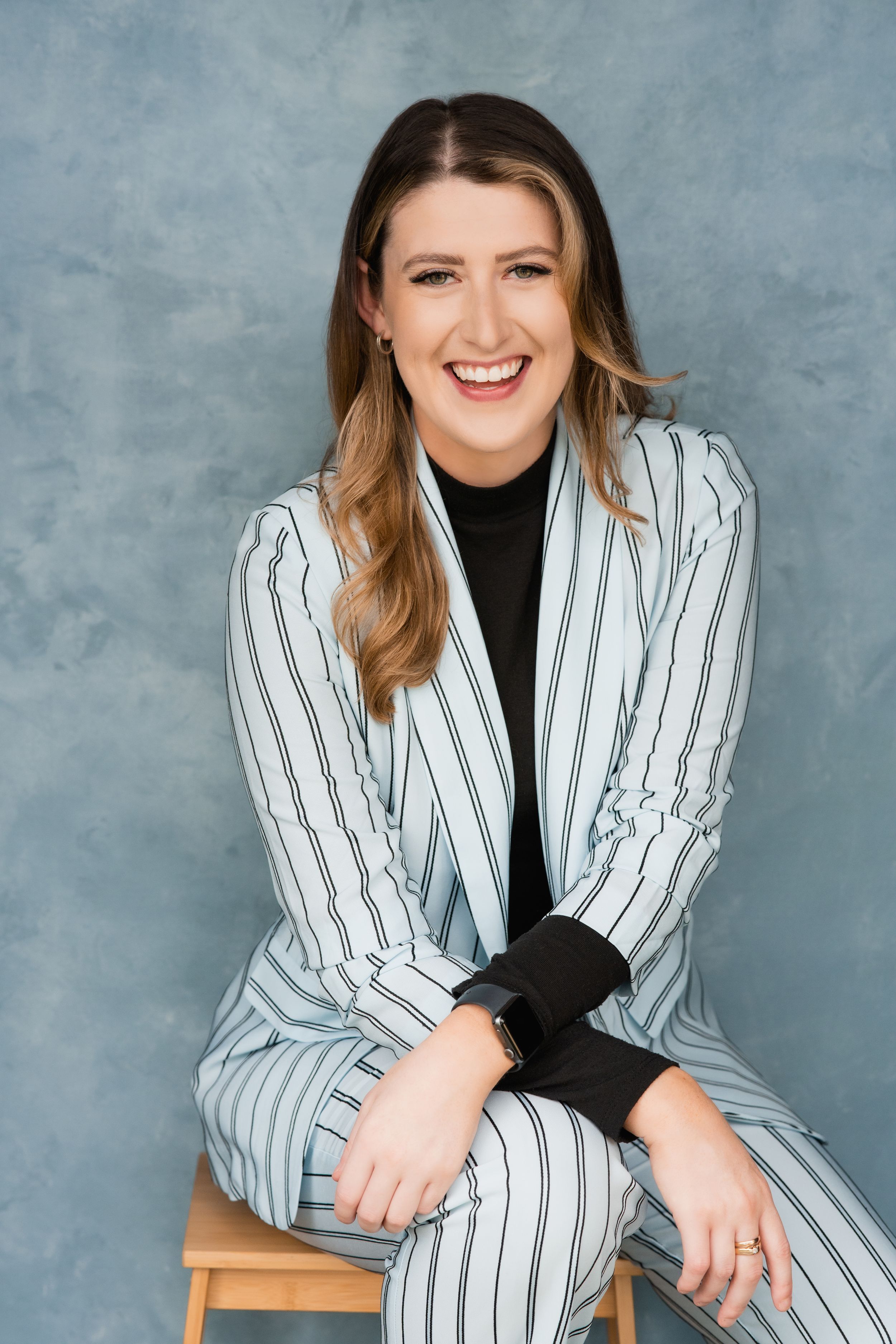 Professional portrait featuring a person in a striped white pantsuit and black top sitting on wooden stool against gray backdrop.