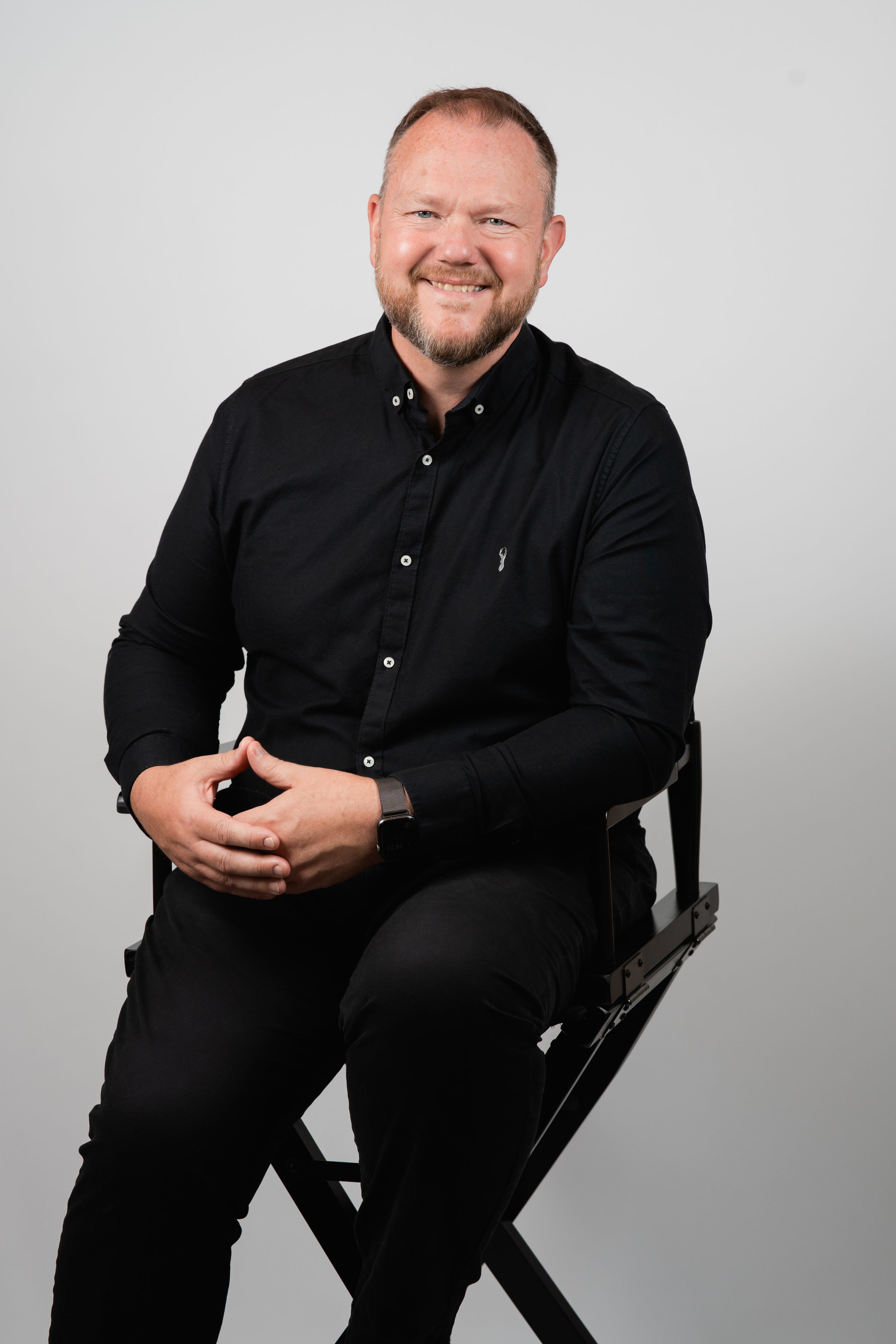 Studio portrait of person in black attire seated on director's chair against light grey backdrop. Headshots in Guildford, Surrey.