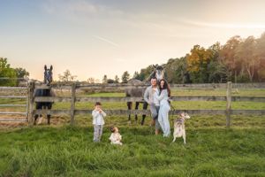 A family stands in front of a wooden fence at sunset on a farm with a horse and dogs in the background.