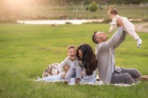 A family sits together on a picnic blanket in a sunny park, enjoying quality time outdoors as a baby is lifted playfully.