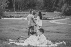 Two children lounging in a grassy field while a couple stands together in the background in black and white.