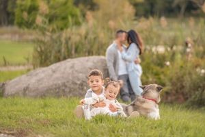 Two young children and a dog relax in a grassy field with people in the background on a sunny day.