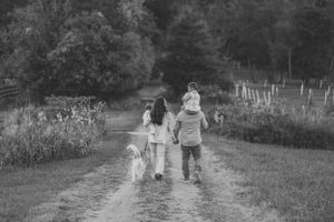 A family walks together with their dog down a dirt path in black and white, surrounded by rural scenery.