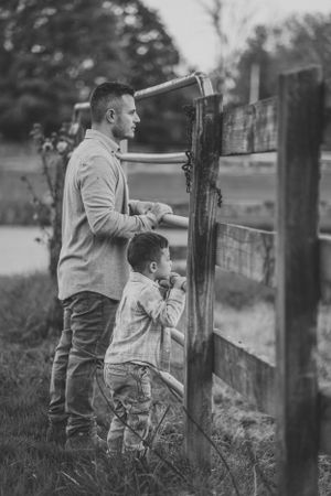 Black and white portrait of a parent and child standing together at a rustic wooden fence in the countryside.