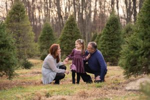 Three people crouching together at a Christmas tree farm surrounded by evergreen trees on a winter afternoon.