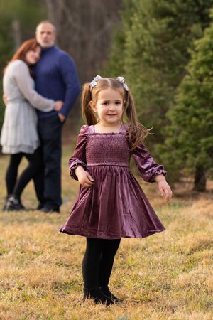Young child in a purple velvet dress and black leggings poses outdoors with nature backdrop.