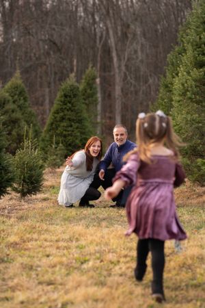 A child in a purple dress runs towards parents crouching by evergreen trees on a winter day.