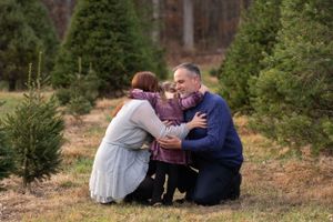 A family shares an intimate moment together while crouched among evergreen trees at a Christmas tree farm.