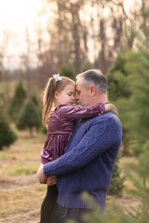 A tender moment shared between a child in a purple dress and father in a blue sweater at a Christmas tree farm at dusk.