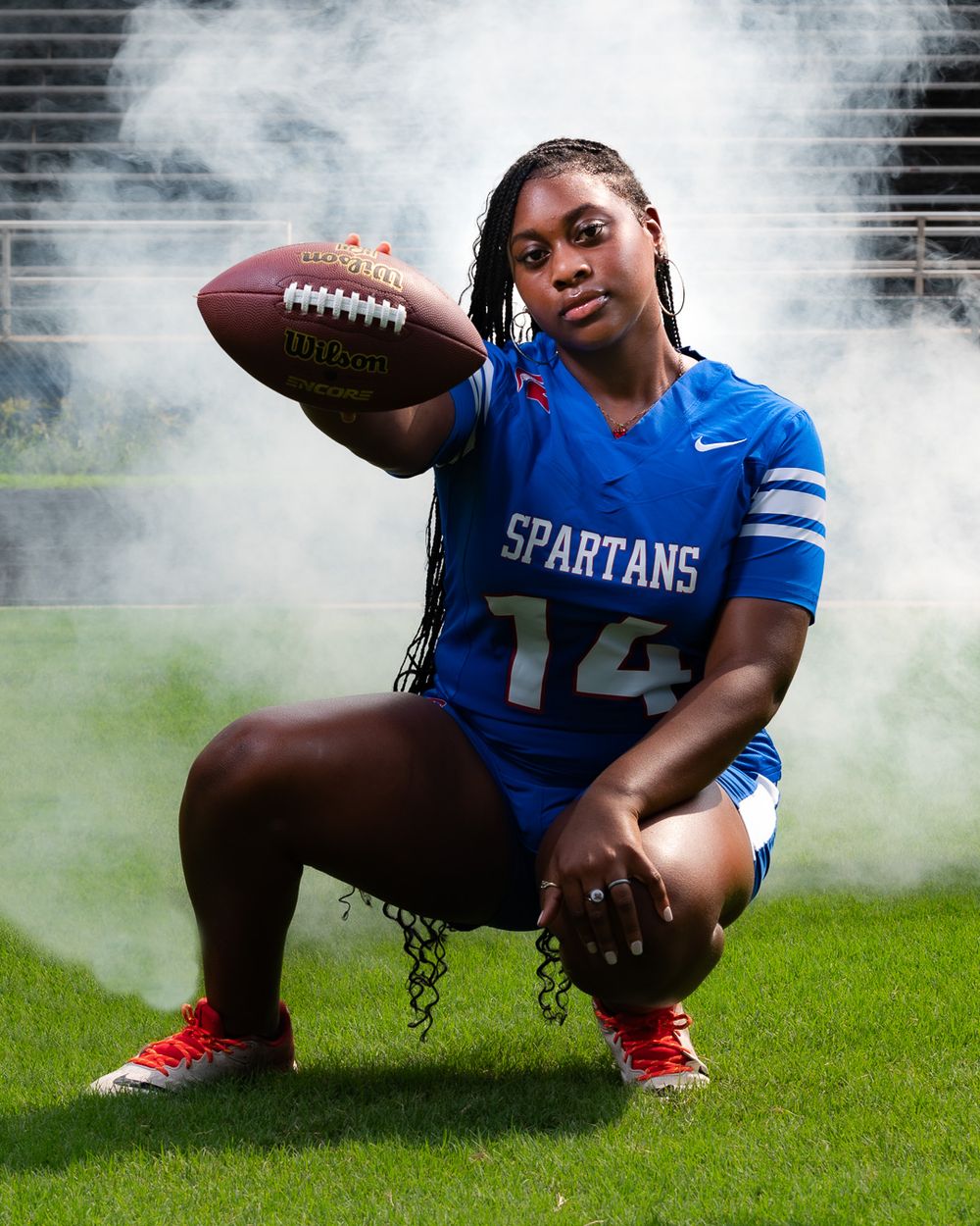 Athlete in Spartans uniform crouches with football on field surrounded by smoke.