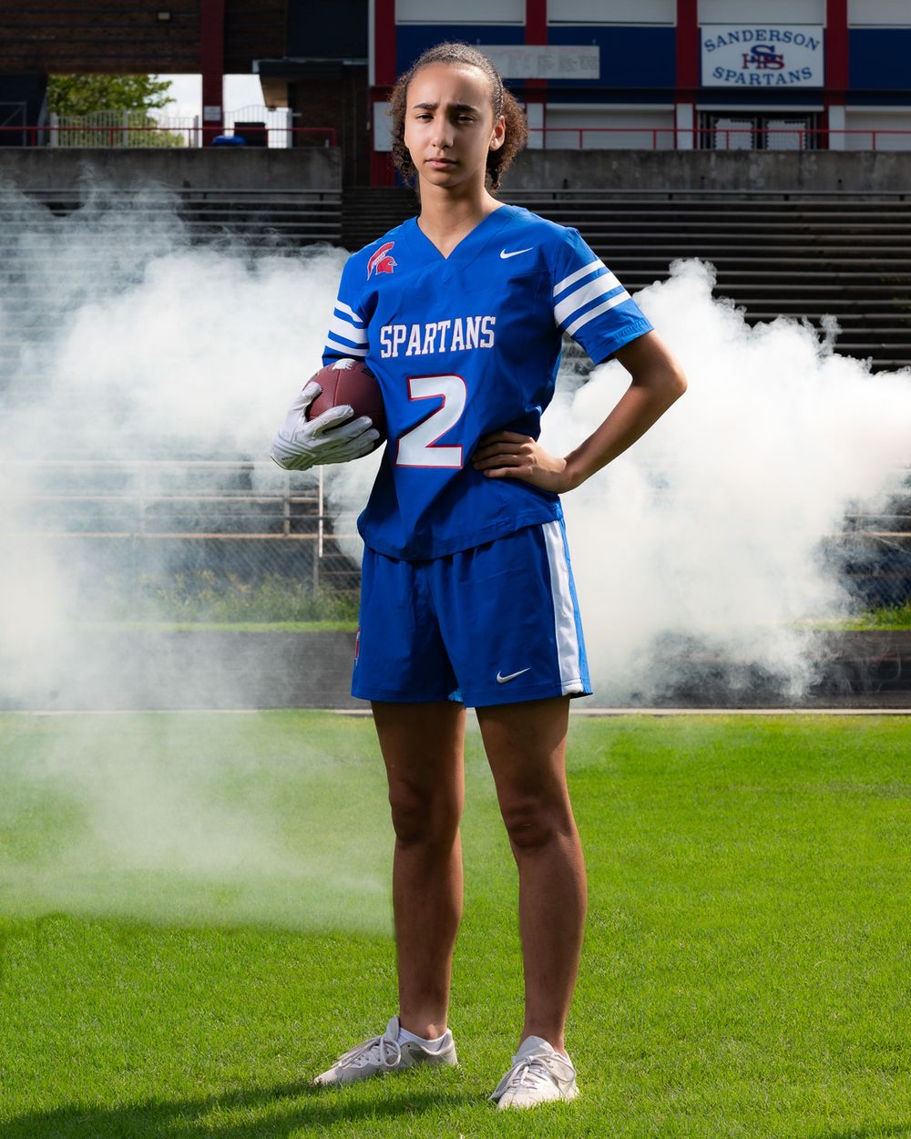 High school athlete poses in blue Spartans jersey on football field with smoke effects in background.