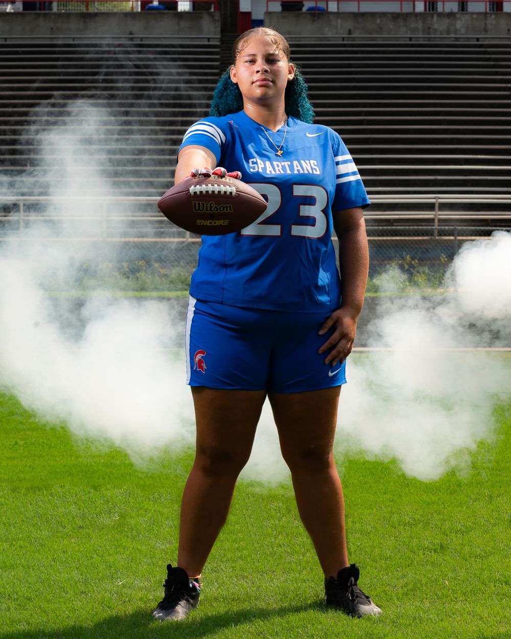 Football player in blue Spartans jersey poses with ball amid smoke effects.