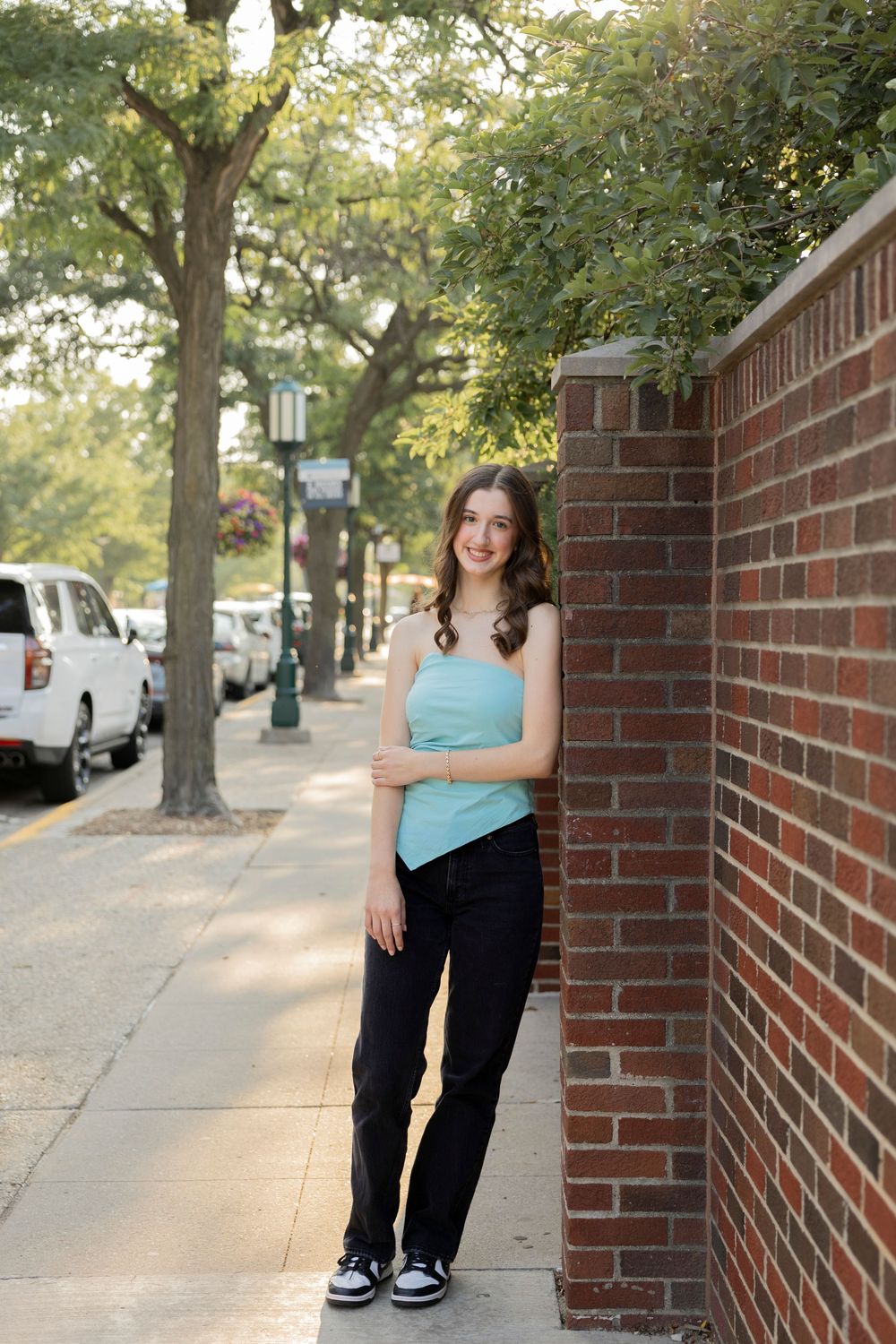 teenage girl posing for her senior photo taken against brick wall with trees and sidewalk in the background in downtown Birmingham michigan