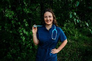 Healthcare professional in blue scrubs poses outdoors against green foliage background.