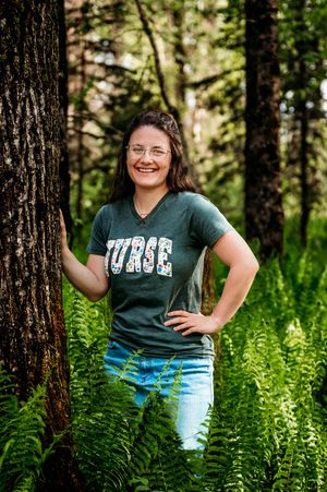 A person wearing a green t-shirt poses casually in a forest setting with lush ferns and pine trees in the background.