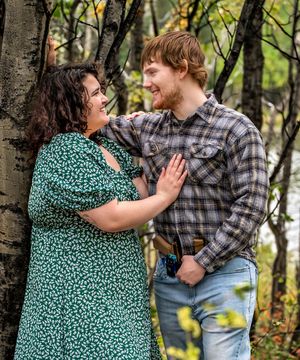 A couple shares a playful moment together while standing in a wooded area on an autumn day.