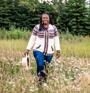 Person in a white patterned sweater sits joyfully in a field of wildflowers on a sunny day.