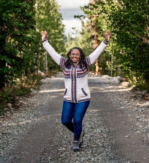 A person in a patterned sweater and jeans jumps joyfully on a gravel path surrounded by greenery.