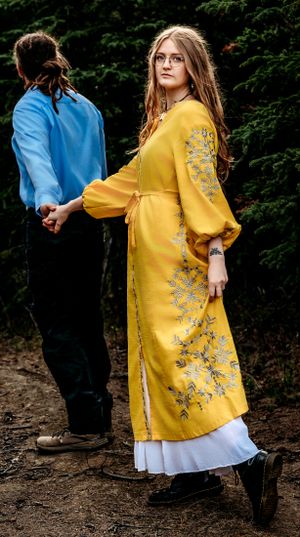 Woman in ornate yellow dress standing on forest path with floral embroidery details.