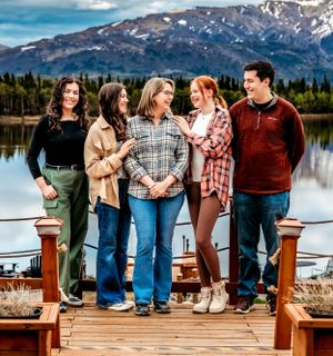 Group standing on wooden deck with snow-capped mountains and lake in background.