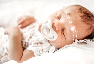 A sleeping baby wearing delicate lace clothing and a pearl headband rests peacefully on white bedding.