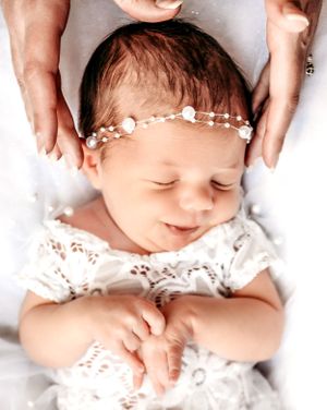 A newborn baby smiles while wearing a delicate pearl headband and white lace outfit.