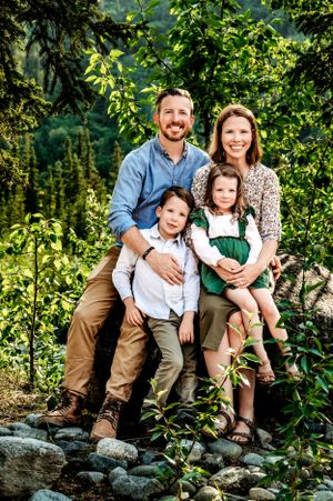 A family of four poses together on rocky terrain surrounded by lush green foliage in an outdoor nature setting.