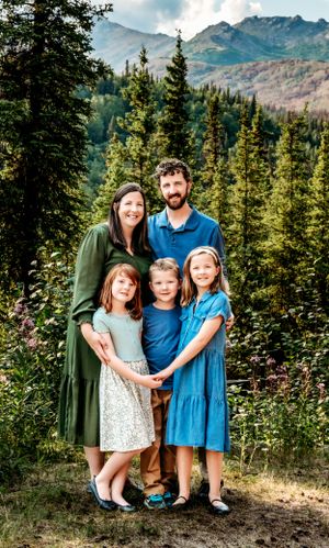 A family group poses together against a backdrop of mountains and evergreen trees in a scenic wilderness setting.